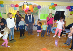 Rodzice z dziećmi tańczą w kole na sali udekorowanej balonami. Na sali są kolorowe światła.